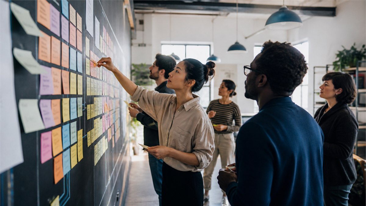 Energetic documentary photograph of a team physically redesigning a colourful workflow together, moving tasks and roles around a room instead of watching a lecture, no text